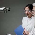 Dois pesquisadores de jaleco e óculos de proteção analisam um braço robótico em um laboratório.