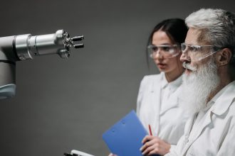 Dois pesquisadores de jaleco e óculos de proteção analisam um braço robótico em um laboratório.
