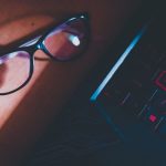 Óculos sobre uma mesa iluminada, ao lado do teclado de um laptop.