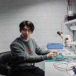 Técnico trabalha com componentes eletrônicos e um microscópio em um laboratório de desenvolvimento de microchips.