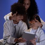Mãe cobre os olhos de duas crianças enquanto elas seguram um tablet, simbolizando o controle parental na internet.
